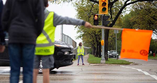 Image showing the back of a school patroller in the foreground with an orange flag extended and another school patroller in the background with arms extended.