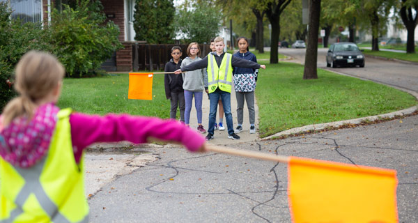 Image showing a school patroller across an intersection with both arms extended and holding back a group of students until it is safe to cross.