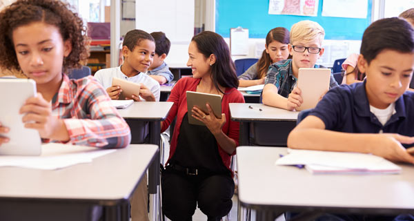 Image showing a classroom full of students at their desks, with a smiling female teacher kneeling beside a students’ desk and talking.